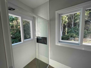 A bathroom with two windows, a glass shower enclosure, and a view of a forested area.