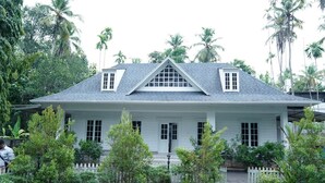 Uma casa de estilo colonial branca com telhado cinzento, rodeada por vegetação exuberante e palmeiras.