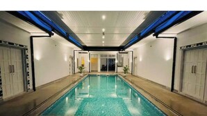 Uma piscina interior retangular com água cristalina, rodeada por paredes com portas e janelas, e iluminação azul no teto.