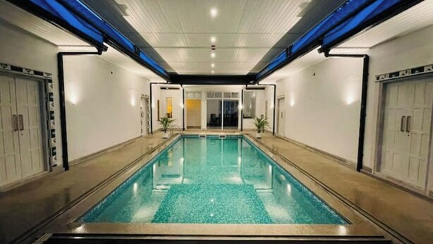 Et rektangulært indendørs swimmingpool med klart vand, omgivet af vægge med døre og vinduer, samt blå belysning i loftet.