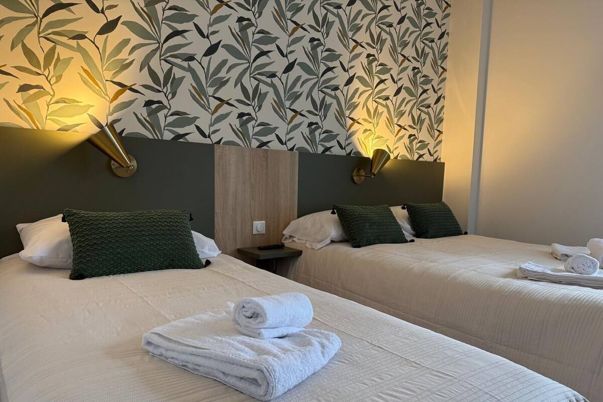Habitación de hotel con dos camas, cada una con una almohada verde y una manta blanca. Hay dos lámparas fijadas a la pared a cada lado de la cama, y una toalla doblada sobre la cama.