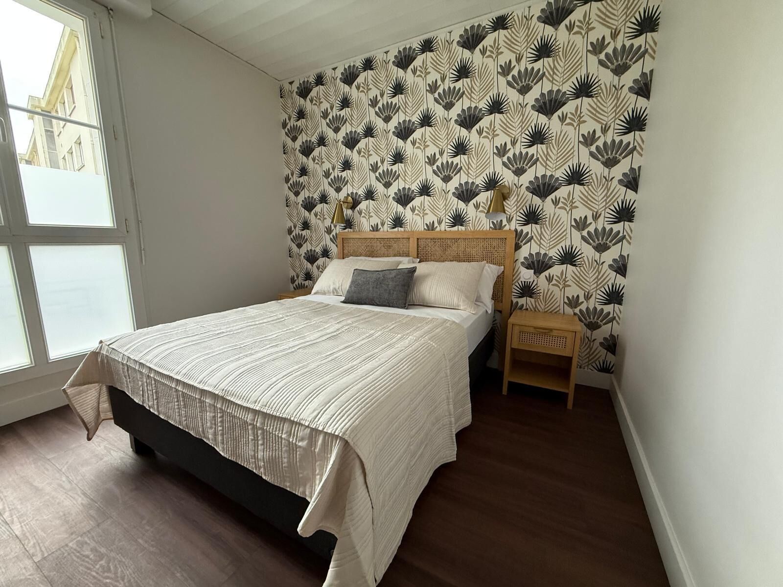 A bedroom with a large bed, a nightstand, a window, and a wall with a floral pattern.