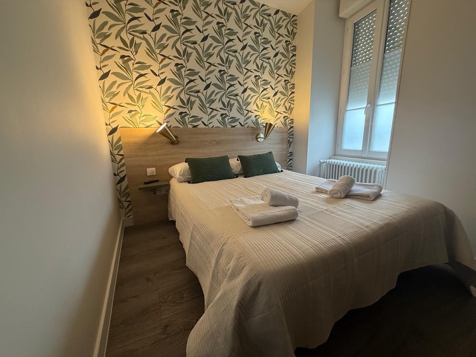 A neatly made bed with white linens and rolled towels, a bedside table with a lamp, and a wall with a leafy pattern.