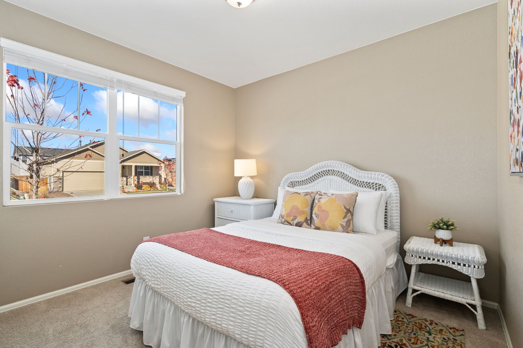 A bedroom with a large window, a bed with a white headboard, a bedside table with a lamp, and a small white side table with a plant.