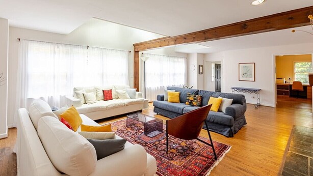 Een ruime woonkamer met een witte bank, een grijze bank en een rode fauteuil. Er ligt een houten vloer en er is een groot raam met gordijnen.