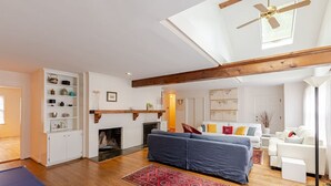 Een ruime woonkamer met een open haard, een blauwe bank, een witte bank en een plafondventilator.