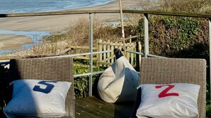 Un balcó davant del mar amb cadires i coixins de vimet, una bandera i vista a la platja i l'oceà.