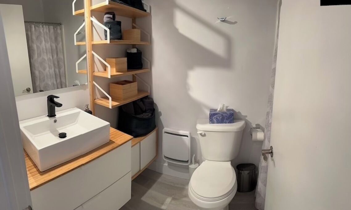 A compact bathroom with a white sink, a toilet, and a mirror. There are wooden shelves above the sink and a small trash bin next to the toilet.