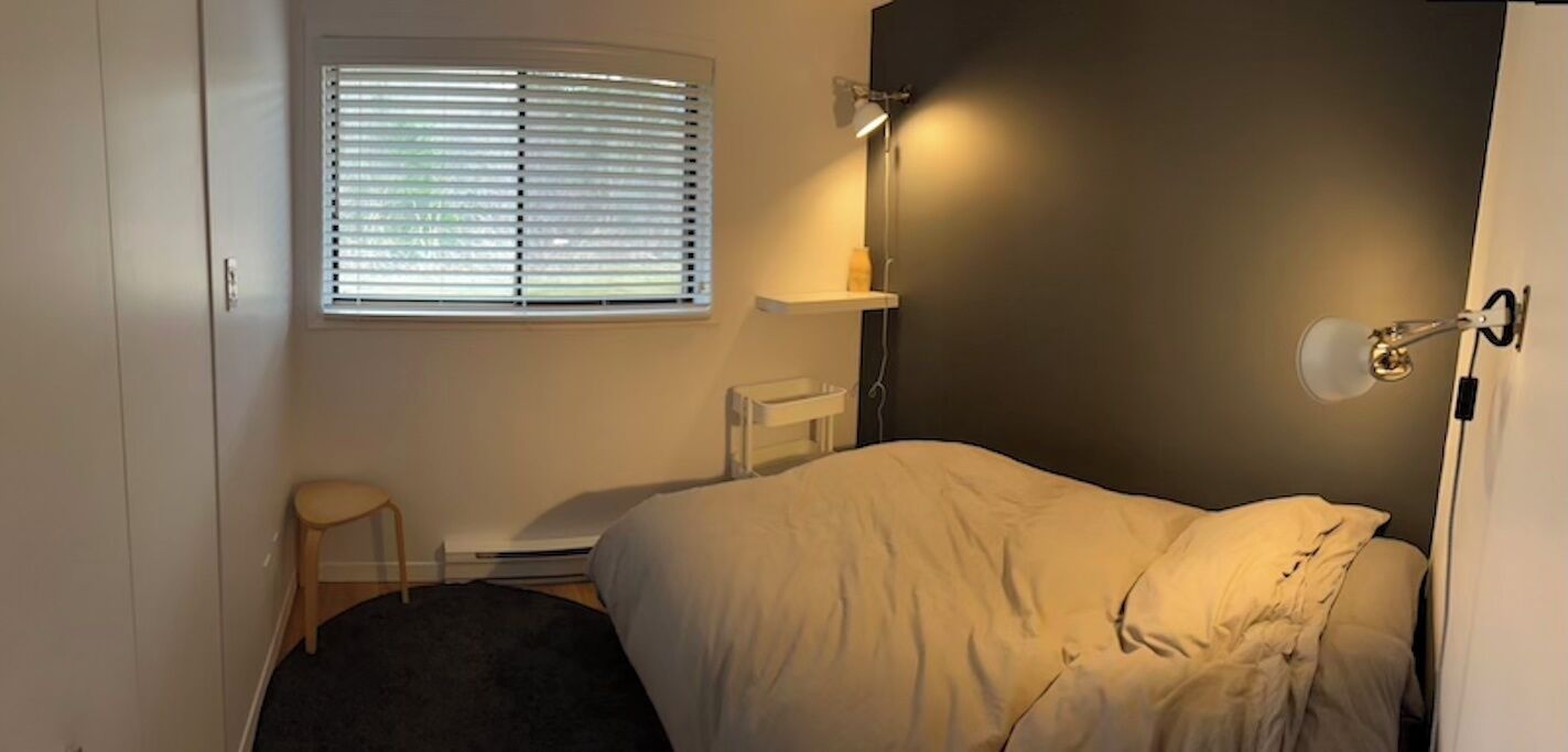 A bedroom with a bed, a desk, a chair, a window with blinds, and a wall-mounted lamp.