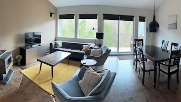 Een moderne woonkamer met een bank, eettafel en stoelen. Er is een open haard, een televisie en grote ramen.