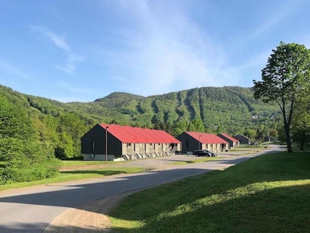 A row of red-roofed buildings with a road leading to them, surrounded by green hills.