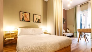 A bedroom with a large bed, a wooden nightstand, a desk with a chair, and two wall-mounted paintings.