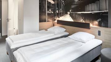 A hotel room with two single beds, a wooden headboard, a large window with a view of a train track, and a door to the left.
