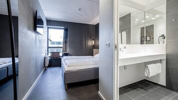 A modern hotel room with a bed, a desk, a chair, a large window, and a bathroom with a mirror and sink.