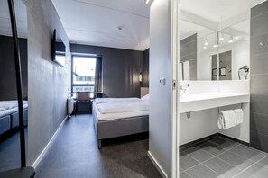 Ein modernes Hotelzimmer mit Bett, Schreibtisch, Stuhl, großem Fenster und einem Badezimmer mit Spiegel und Waschbecken.