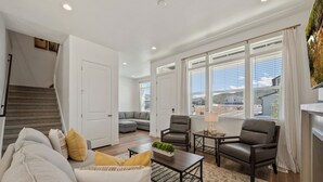 A spacious living room with a sofa, armchairs, a coffee table, and a rug. There is a staircase leading to another room, and large windows with curtains.