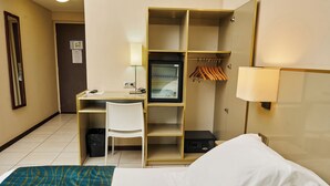 A hotel room with a bed, a desk, a chair, a TV, a coat rack, and a trash bin. - Amazonia de Saint-Laurent (Saint Laurent du Maroni)