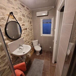 Une salle de bain avec un mur en pierre, un miroir rond, un lavabo, des toilettes et une fenêtre avec des stores bleus.