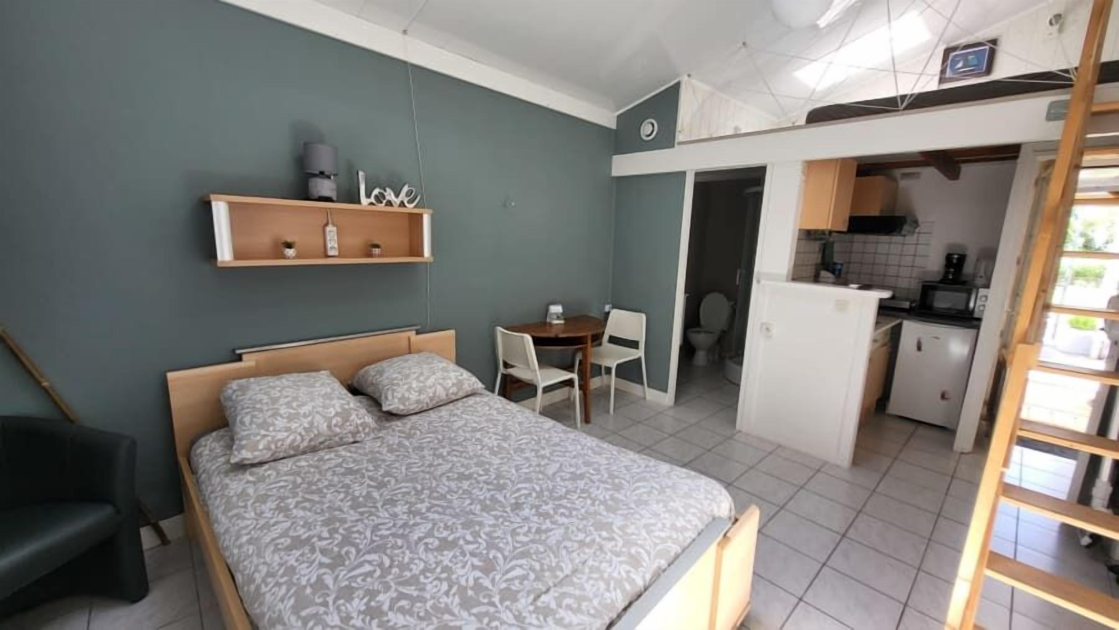 Ein Schlafzimmer mit Bett, Schreibtisch und Stuhl. Anliegend befindet sich eine Küchenzeile.