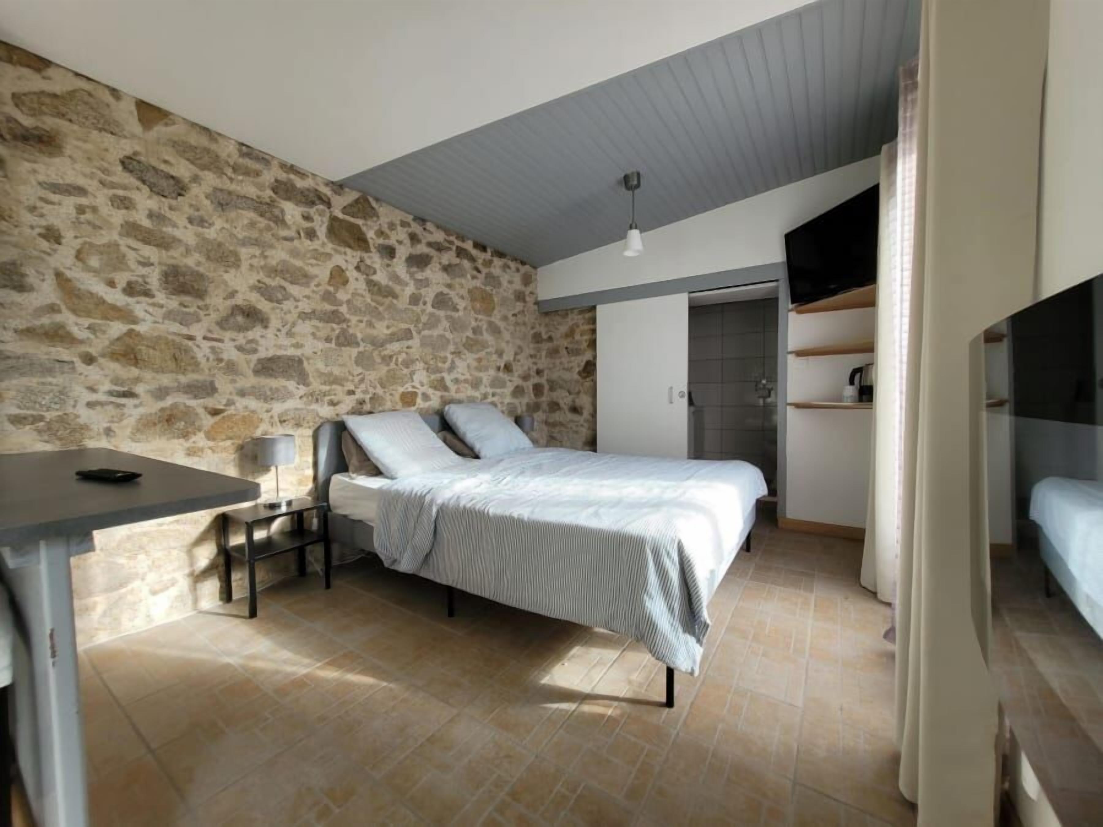 Een slaapkamer met een stenen muur, een bed met een witte spreideken, een klein tafeltje met een lamp en een inbouwkast.