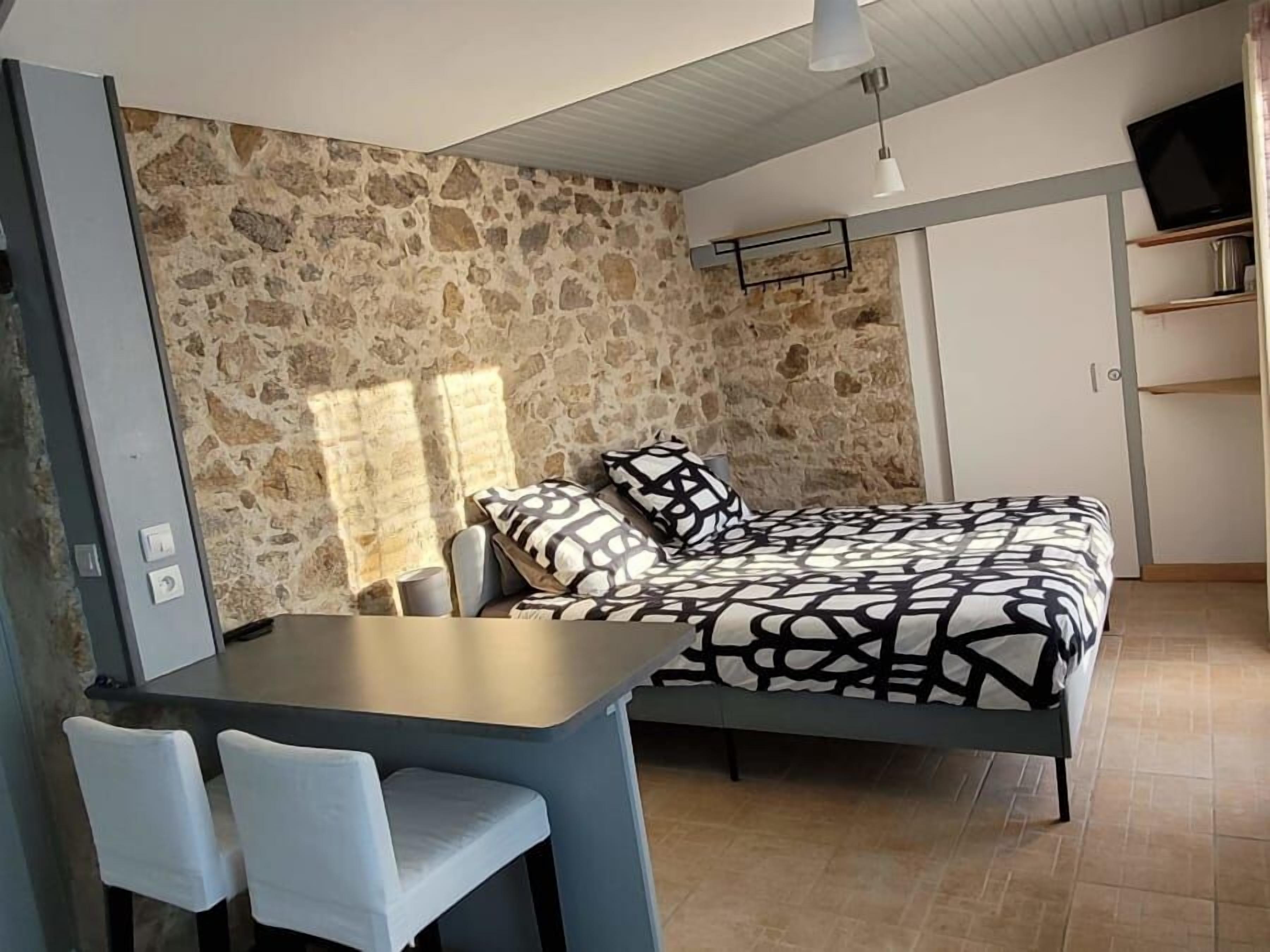 Een slaapkamer met een stenen muur, een bed met beddengoed met een patroon, een bureau met stoelen en een televisie op een plank.