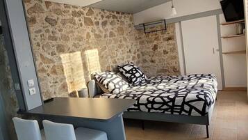 Een slaapkamer met een stenen muur, een bed met beddengoed met een patroon, een bureau met stoelen en een televisie op een plank.
