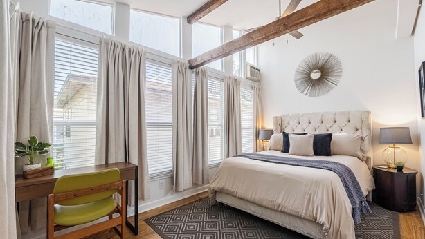 A bedroom with a large bed, a desk, and a chair. There are curtains on the windows, a wall clock, and a small table with a lamp. - The Loft at Raintree (Lakeland)