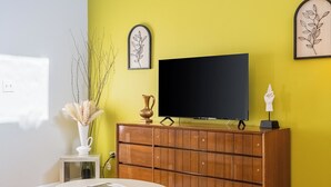 A living room with a yellow wall, a wooden cabinet, a flat-screen TV, a white vase with pampas grass, and a small table with a magazine. - The Loft at Raintree (Lakeland)