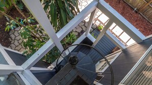 A spiral staircase with a handrail, surrounded by a white railing and wooden steps, leading to a covered outdoor area with plants and a stone wall. - The Loft at Raintree (Lakeland)