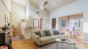 A spacious living room with a sofa, coffee table, and a patterned rug. A kitchen area with a refrigerator and stove is visible in the background. - The Loft at Raintree (Lakeland)