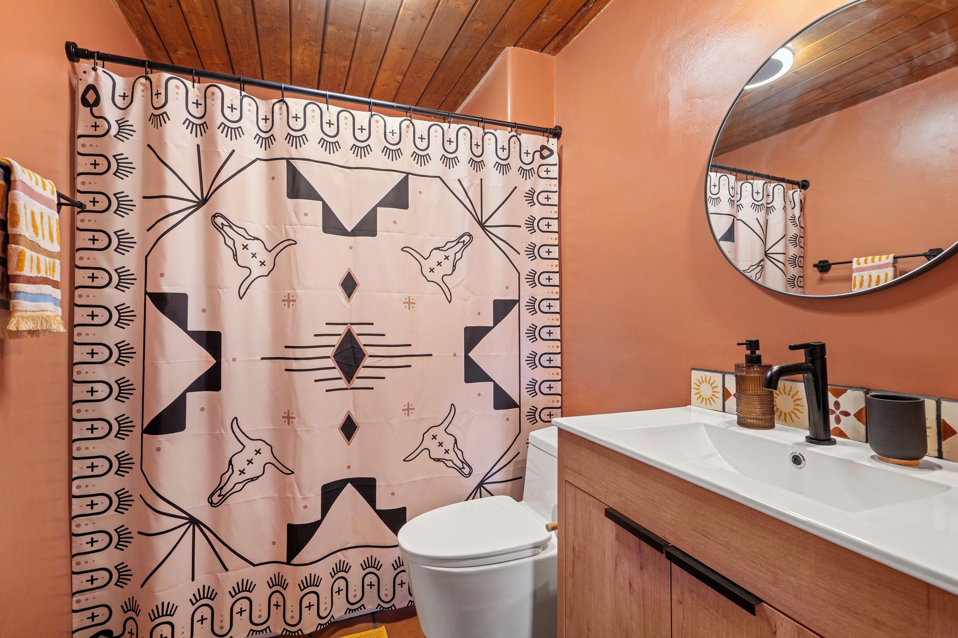 Un bagno con tenda da doccia a motivi geometrici, un water bianco, un mobile bagno in legno con lavandino e uno specchio sopra il lavandino.