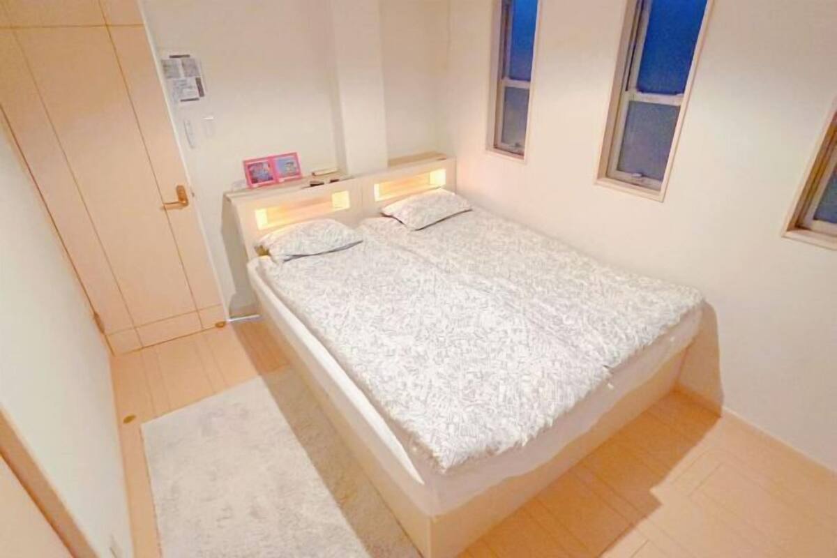 Un llit individual amb un edredó blanc, una taula de nit amb una làmpada i una finestra amb vista al cel.
