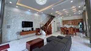 A spacious living area with a large sofa, a TV mounted on the wall, a staircase leading to the upper floor, and a kitchen area visible in the background. - Sunrise Villa (Phu Quoc Island)
