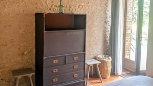 A bedroom with a bed, a dresser, a small stool, a wicker basket, and a window with curtains. - Gîtes du Martroy (Nargis)