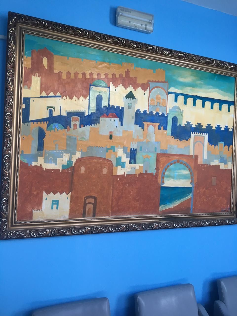 Un cuadro enmarco muestra un paisaje urbano costero con un puente y torres, exhibido sobre una pared azul.