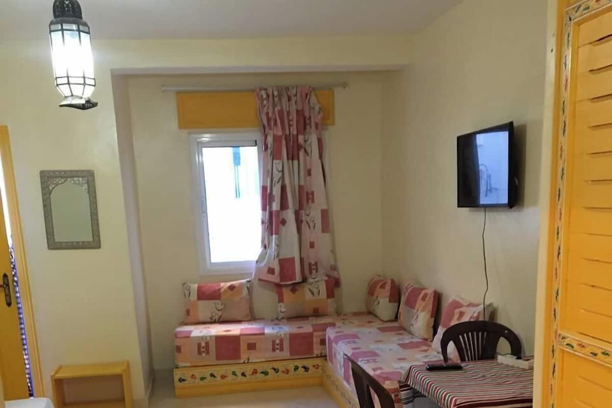 A cozy room with a bed, a small table, a chair, a TV, and a window with curtains.