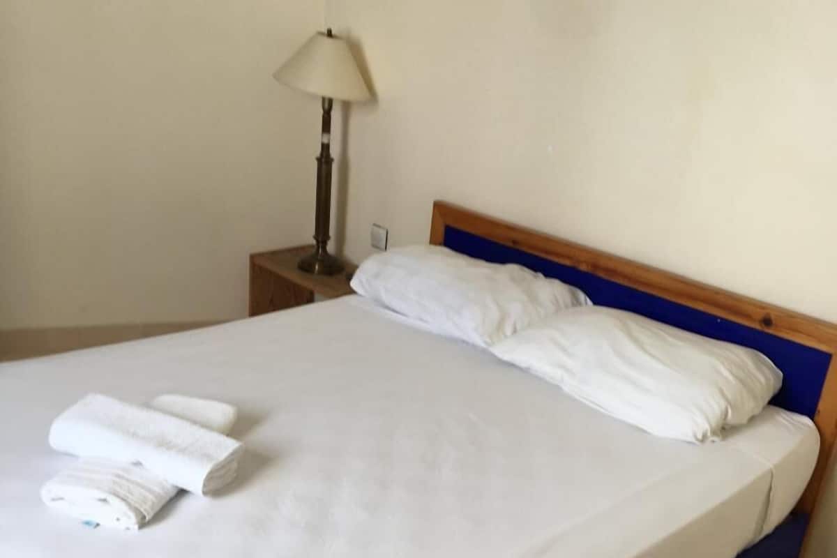 A neatly made bed with white linens and two rolled towels, a wooden headboard, a bedside table with a lamp, and a plain wall.