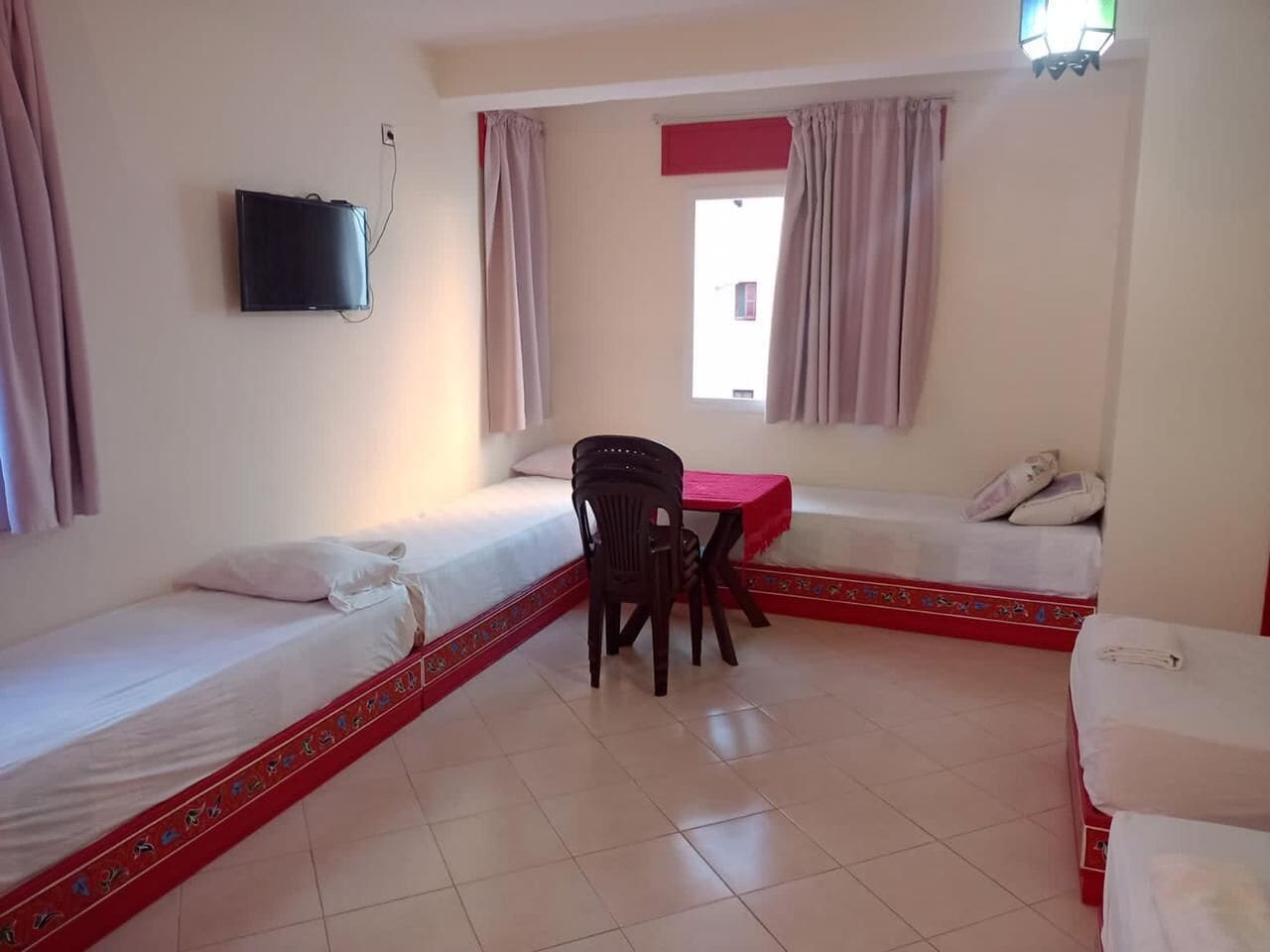 Una habitación con dos camas individuales, una mesita con una silla, un televisor en la pared y una ventana con cortinas.