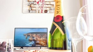 Una bottiglia di champagne Moët & Chandon, una ciotola di frutta secca, un bicchiere d'acqua e un televisore che mostra un paesaggio costiero.