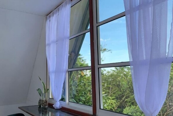 A room with a large window, white curtains, and a view of greenery outside.