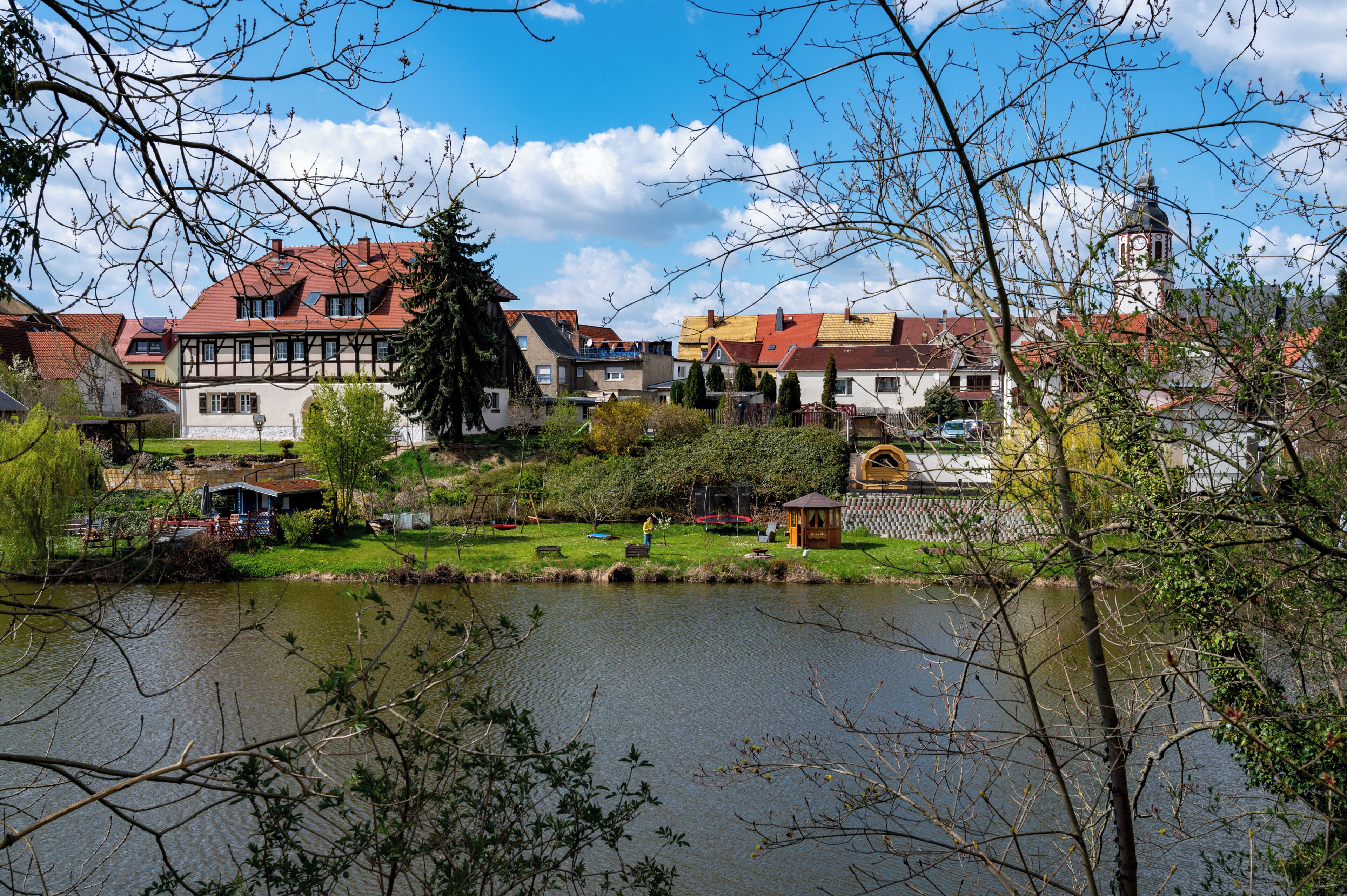 A riverside view of a town with traditional buildings, a clock tower, and a gazebo by the water.