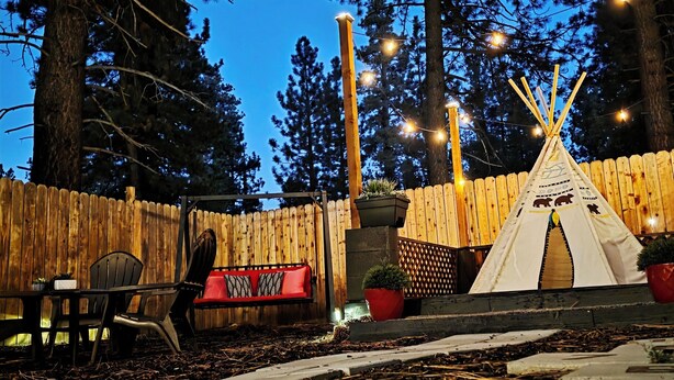 A wooden deck with a red sofa and chairs, a teepee, and string lights against a backdrop of tall trees.