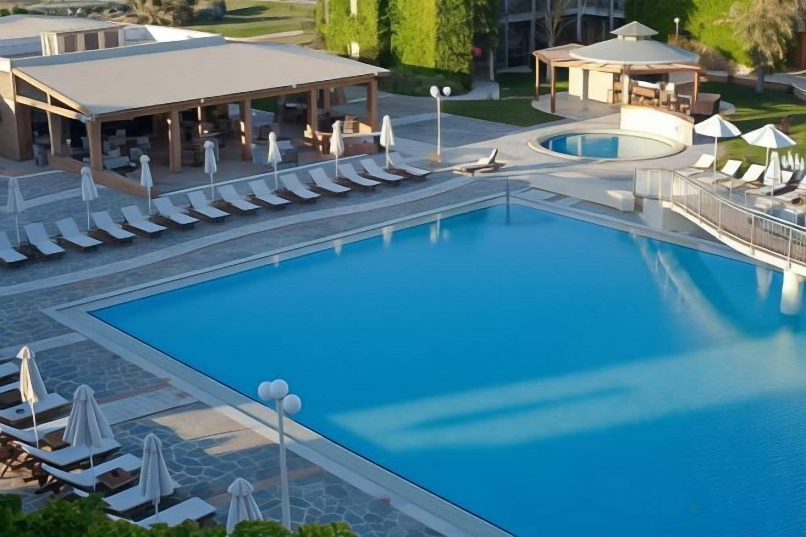 Una gran piscina con tumbonas y sombrillas, rodeada de un área de estar techada y una pequeña piscina circular.
