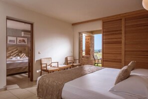 A bedroom with a bed, a seating area with chairs, a wooden headboard, and a view through an open door. - Bella Beach Hotel (Agkisaras)
