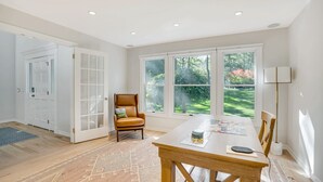 A spacious room with large windows, a wooden dining table, a leather chair, and a floor lamp.