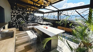 Un espace de restauration extérieur moderne, doté d’un toit métallique aux formes géométriques, d’un bar en béton et d’un mur orné d’œuvres d’art abstraites.