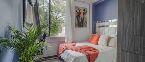 Un dormitorio con cama, ventilador de techo, ventana con cortinas, un cuadro de un elefante y una planta en maceta.