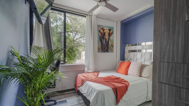 Une chambre à coucher avec un lit, un ventilateur de plafond, une fenêtre avec des rideaux, un tableau représentant un éléphant et une plante en pot.