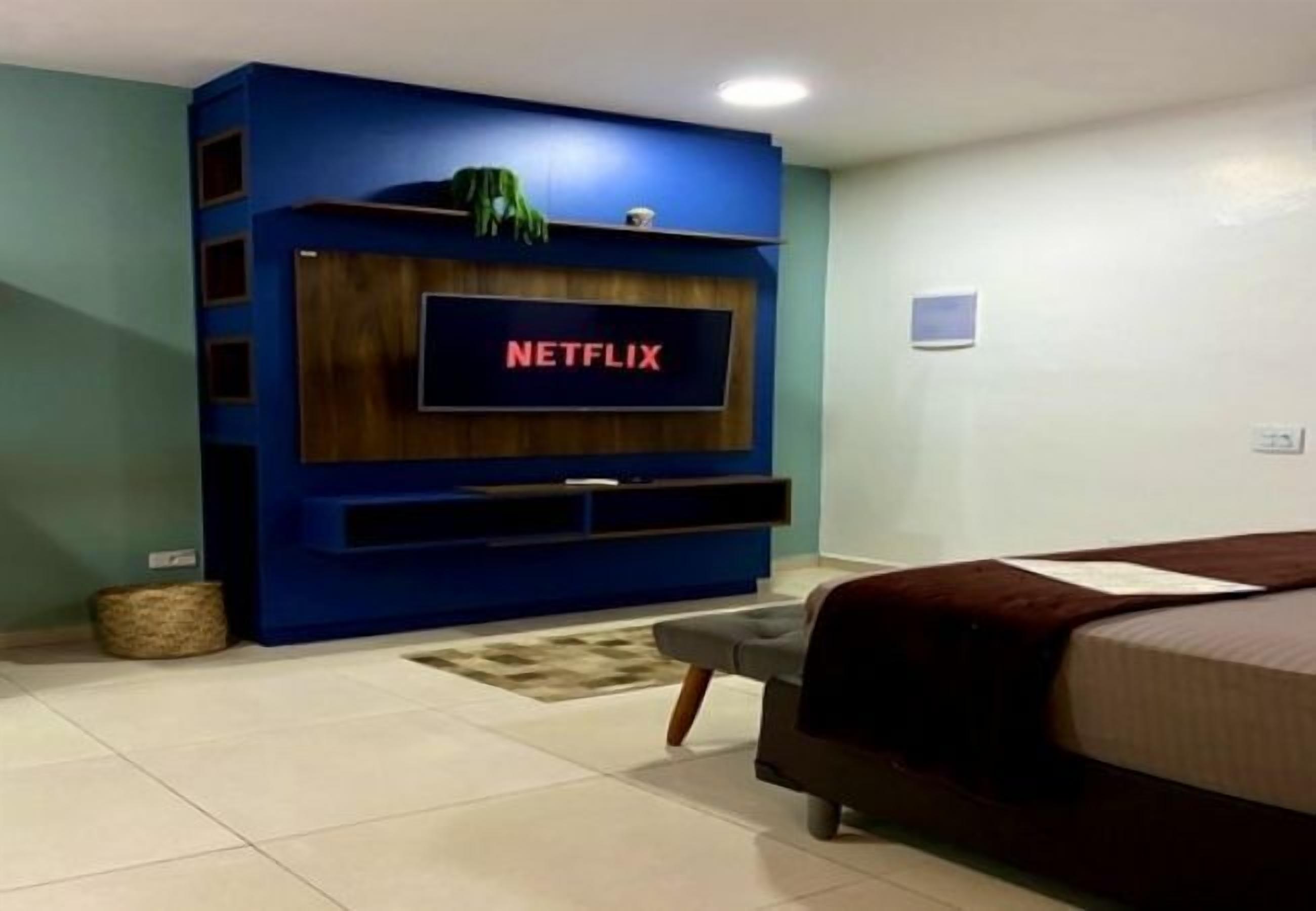 A modern living room with a large flat-screen TV mounted on a blue entertainment unit, a bed with a brown headboard, and a small rug on the tiled floor.
