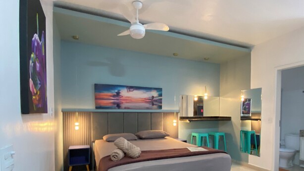 A modern bedroom with a large bed, a ceiling fan, a bench, and a view into the adjacent bathroom. - Mangata Loft (Itanhaém)
